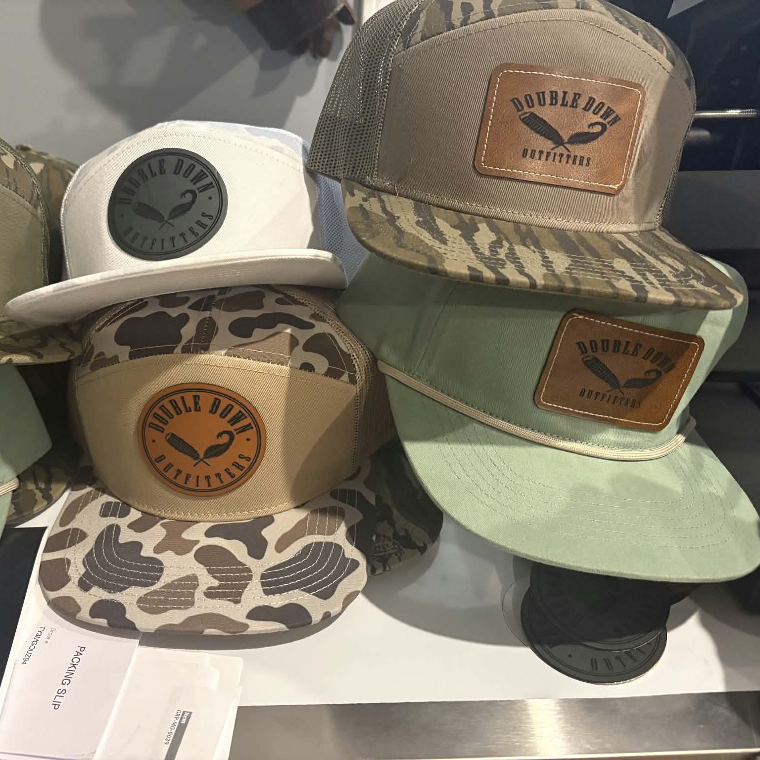 A group of Double Down trucker and flat-brim caps displayed on a retail counter. The collection includes various colorways: olive green and brown camouflage, solid white, light beige, sage green, and taupe with brown leather patches.